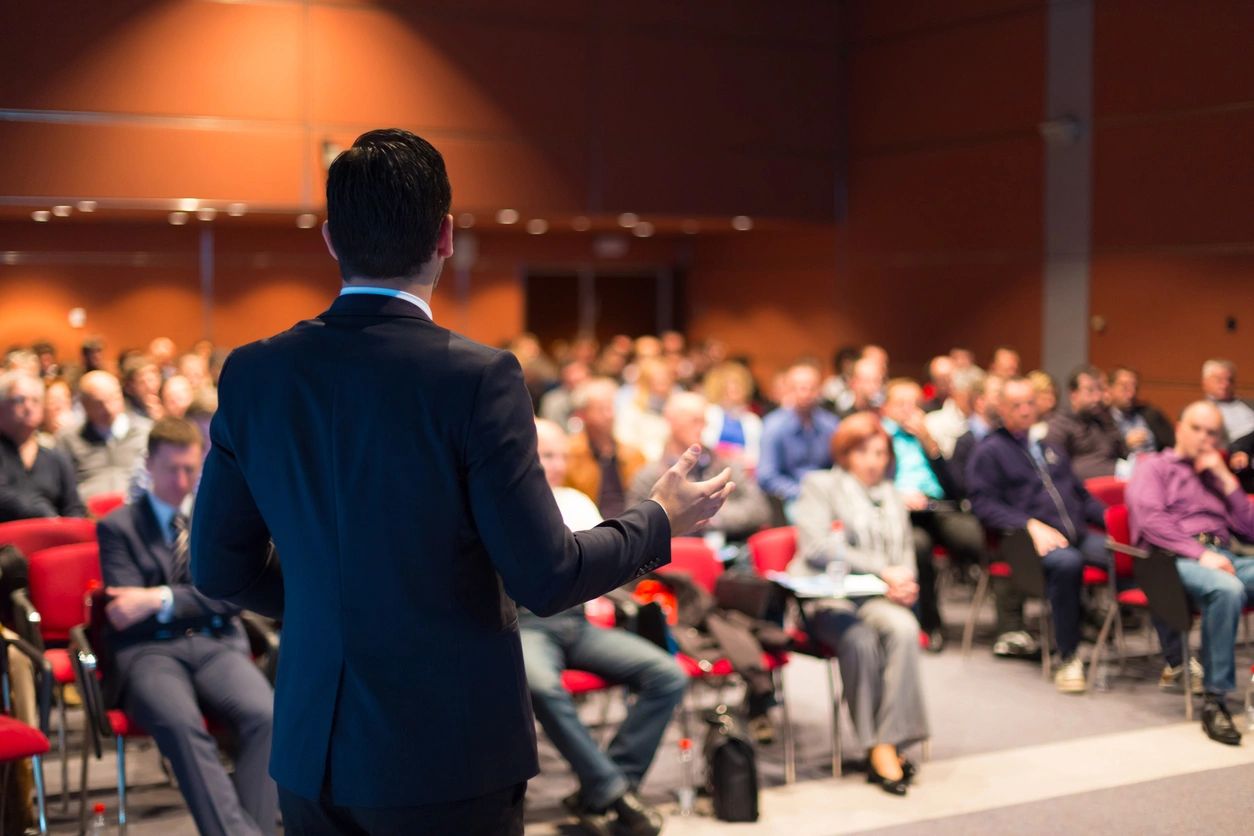 speaker in a public speaking event, facing a crowd gestures open hands as he makes an interesting point