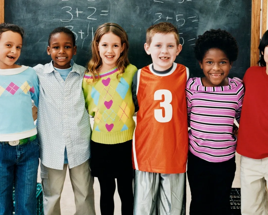 A group of six diverse elementary school children smile for a photo, standing side-by-side in front of a chalkboard.