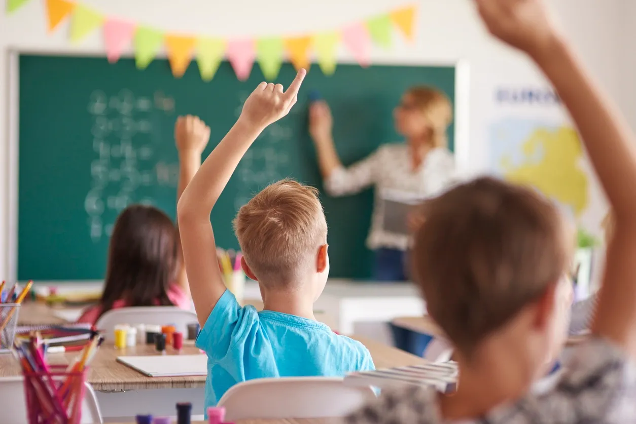 The back of a student raising his hand in a classroom. A teacher writes on a chalkboard in the background.