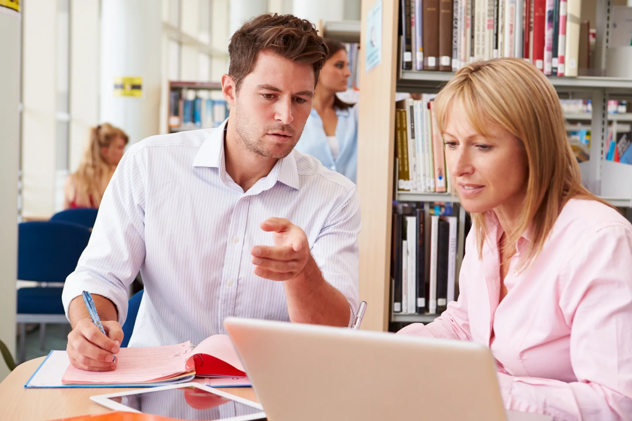 colleagues sitting at a library, with the man pointing and showing his woman colleague on her laptop