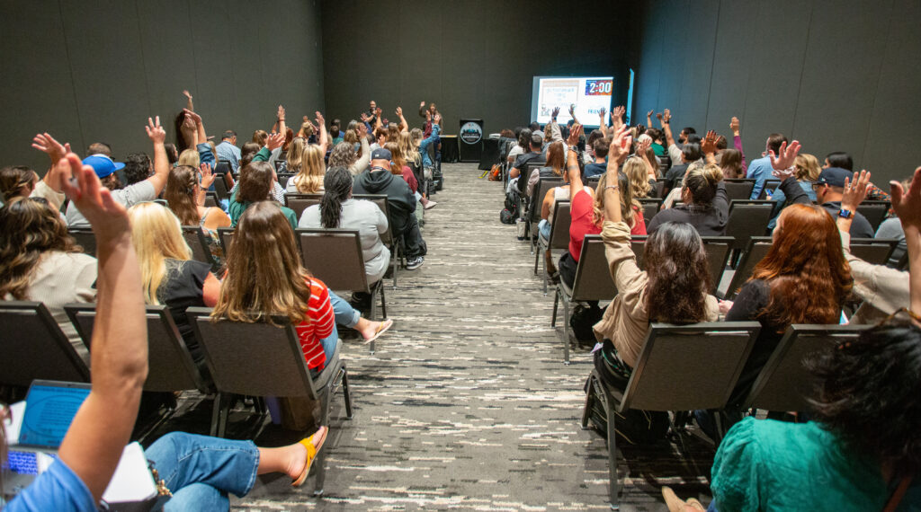 room full of attendees sitting in chairs theater style. Select attendees are raising their hands