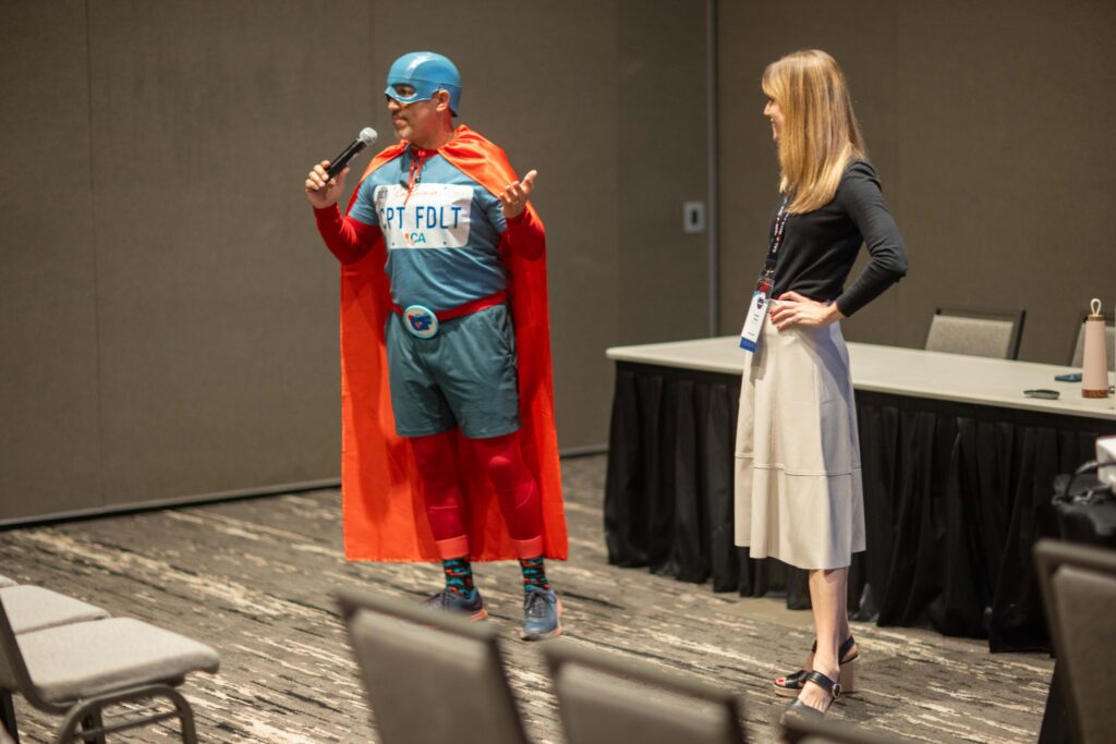 two speakers stand in front of room, one is holding microphone and also is dressed as superhero