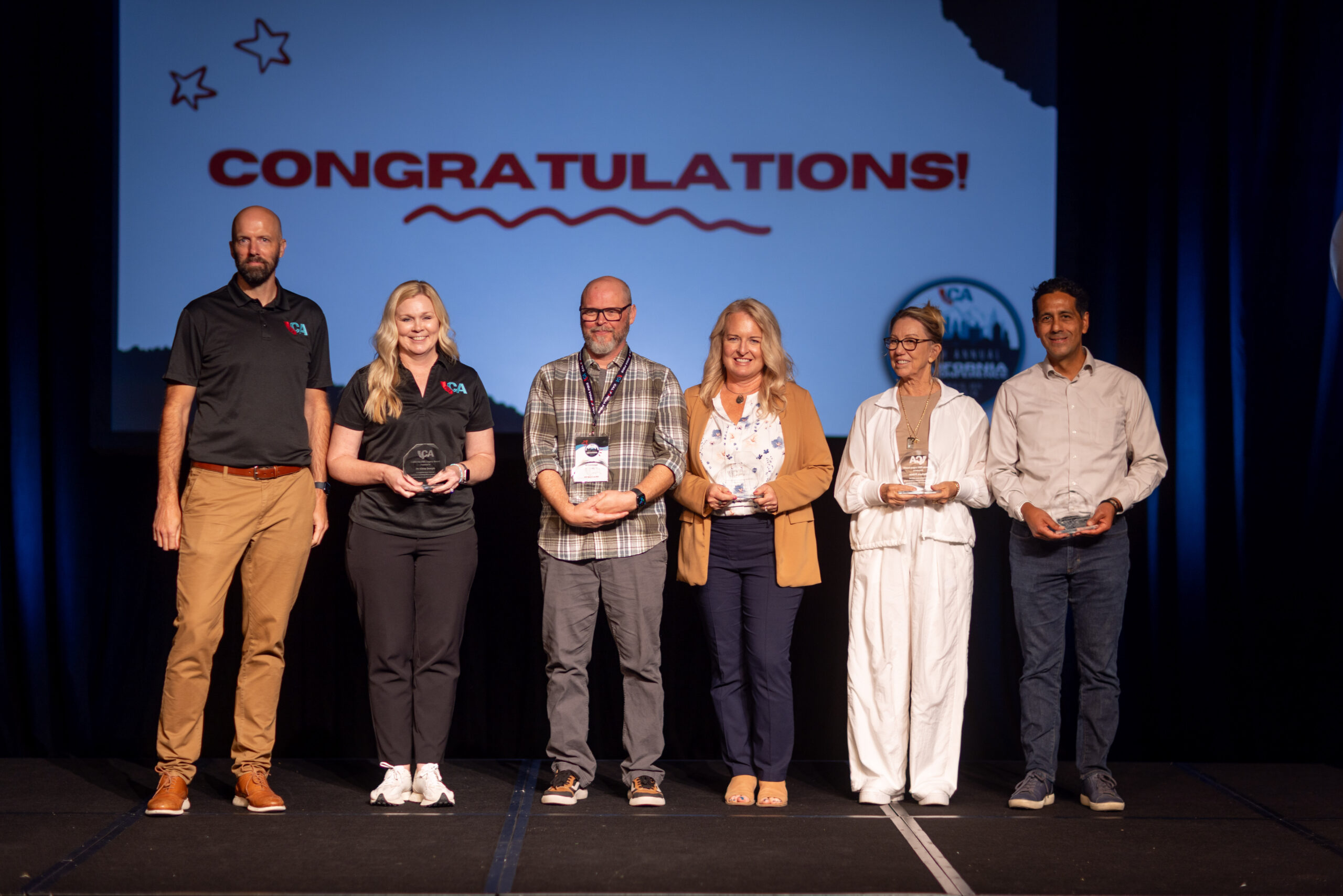 group of 6 people stand on stage in front of screen that reads congratulations. 5 of 6 people hold a glass award