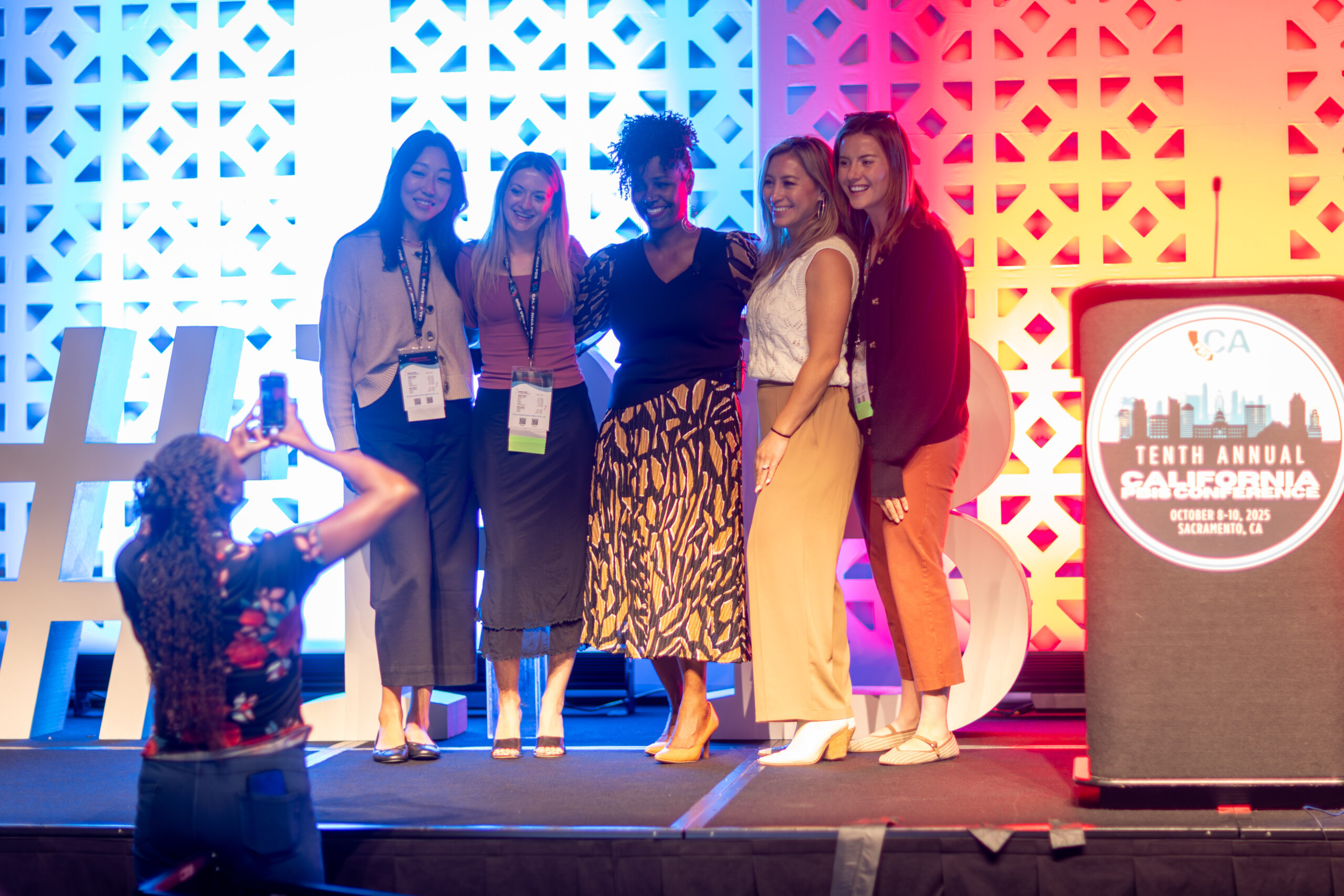 group of 5 women stand on stage smiling while one person stands in front taking photo on cell phone