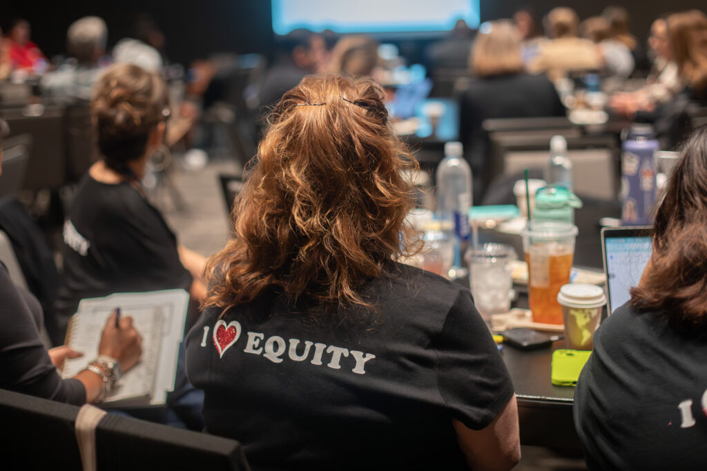 person sits at table facing speaker, back of shirt reads "I heart equity"